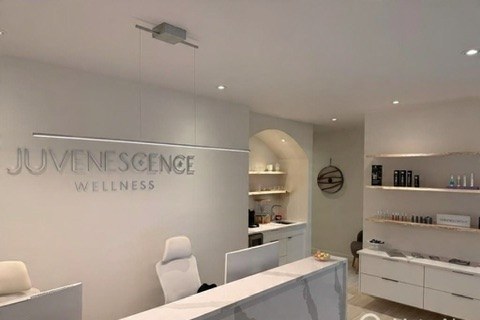 Reception area at Juvenescence Wellness aesthetic clinic in Lake Cowichan, BC — JUVENESCENCE WELLNESS signage on warm-white wall, marble-top desk, curated skincare shelving